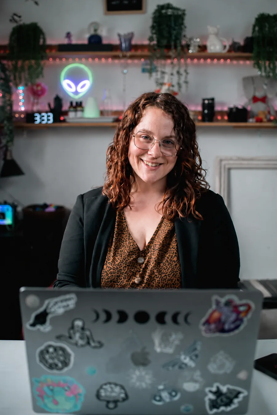 Stéphanie Guilbeault souriante derrière son ordinateur couvert d’autocollants, devant des étagères avec plantes, néon d’extraterrestre et horloge à 3:33.