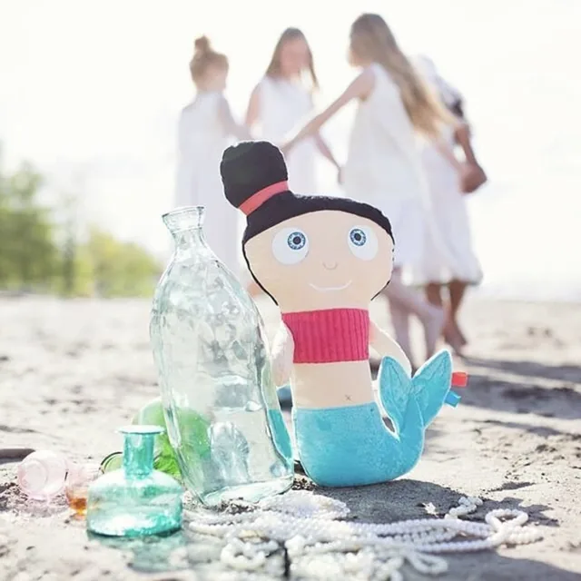 Peluche sirène des TrOnches sur une plage, avec des bouteilles de verre et des perles.